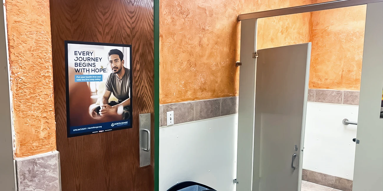 Interior of a bathroom with an orange wall, a wooden door with a poster, and a bathroom stall door. Transcribed Text: EVERY JOURNEY BEGINS WITH HOPE. Put your health first and take the first step today.