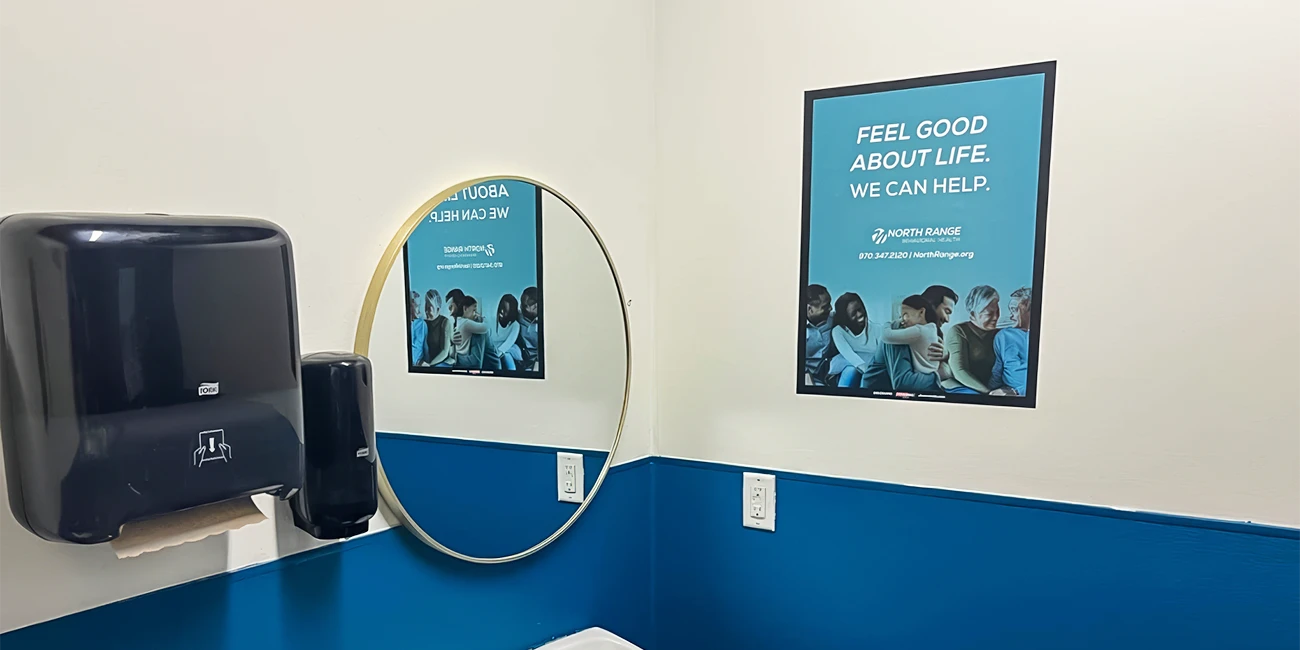 A bathroom with a black paper towel dispenser, a gold-framed round mirror, and a blue poster on the wall. Transcribed Text: FEEL GOOD ABOUT LIFE. WE CAN HELP. North Range Behavioral Health 970.347.2120 | NorthRange.org