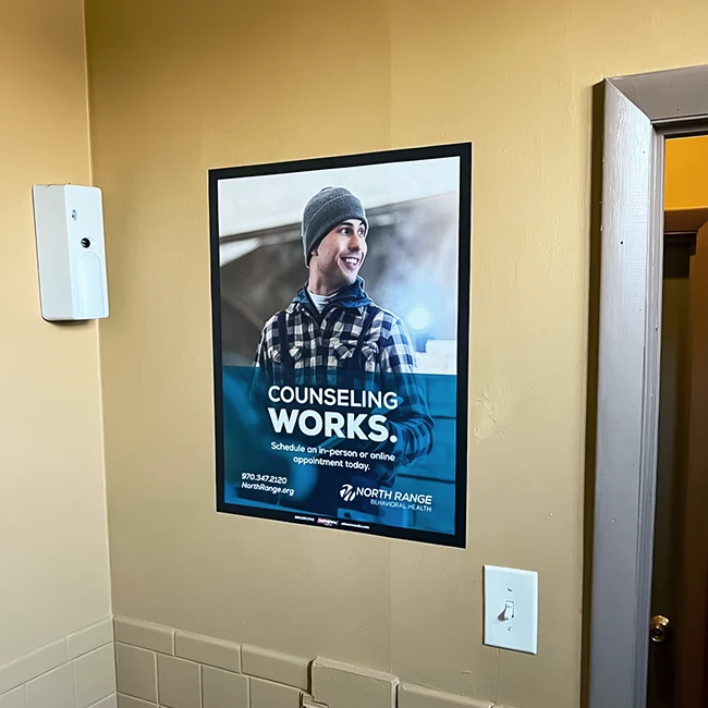 Poster featuring a smiling person in a beanie and a plaid shirt, with text about counseling on a beige wall. Transcribed Text: COUNSELING WORKS. Schedule an in-person or online appointment today. 970-347-2120 NorthRange.org NORTH RANGE BEHAVIORAL HEALTH