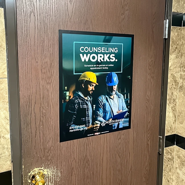 Poster on a wooden door showing two construction workers wearing hard hats. Transcribed Text: COUNSELING WORKS. Schedule an in-person or online appointment today. NORTH RANGE 970-347-2120 NorthRange.org