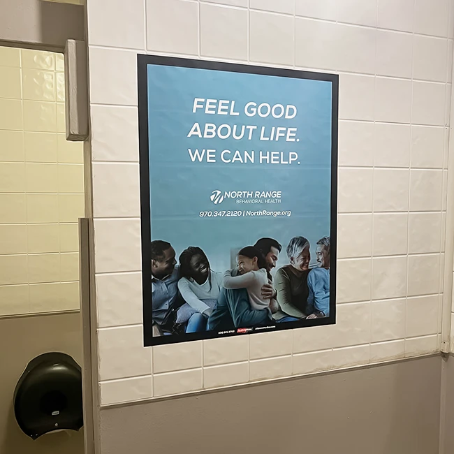 A poster on a tiled wall promoting North Range Behavioral Health with a group of people smiling together. Transcribed Text: FEEL GOOD ABOUT LIFE. WE CAN HELP. NORTH RANGE BEHAVIORAL HEALTH 970.347.2120 | NorthRange.org