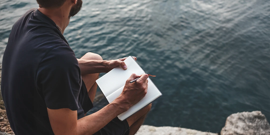 Mental Health Tips Male writing mental health notes in a notebook by the water
