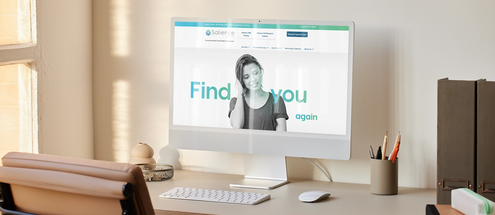 A modern workspace with an Apple desktop computer displaying the "Salience" website. The screen features a greyscaled image of a woman and the text "Find you again" in turquoise. The desk includes a wireless keyboard, mouse, decorative stone, and a holder with pens and pencils.